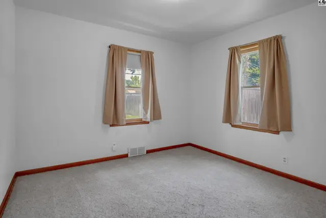 Empty bedroom with neutral carpet, white walls, and two windows with tan curtains.