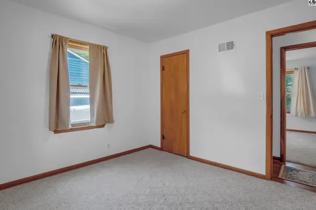 Bright bedroom with neutral carpet, white walls, wood trim, and a doorway to another room.