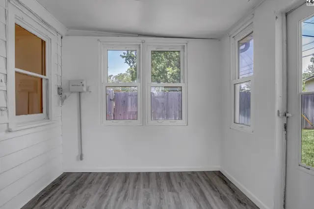 Sunlit room with white paneled walls, wood-look flooring, and large windows overlooking a fenced yard.