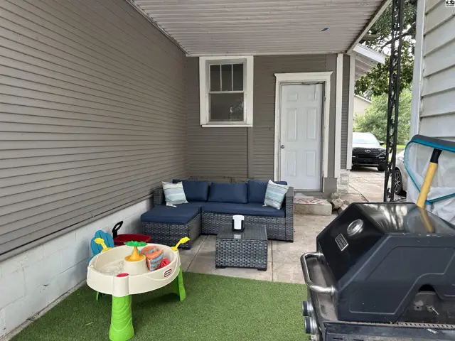 Covered patio with gray siding, wicker furniture, a grill, and a children's play table.