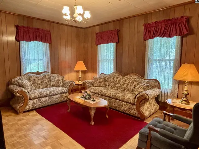 Living room with wood-paneled walls, floral sofas, a red rug, and a vintage chandelier.