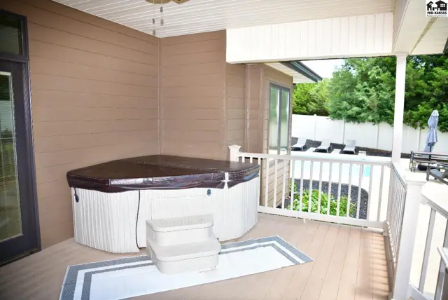 Covered patio with a large hot tub, deck, and view of a swimming pool and trees.