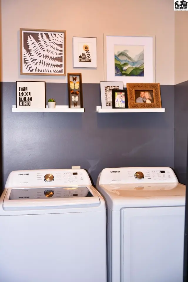 Two white top-load washing machines sit below a wall with two shelves displaying framed art and photos.