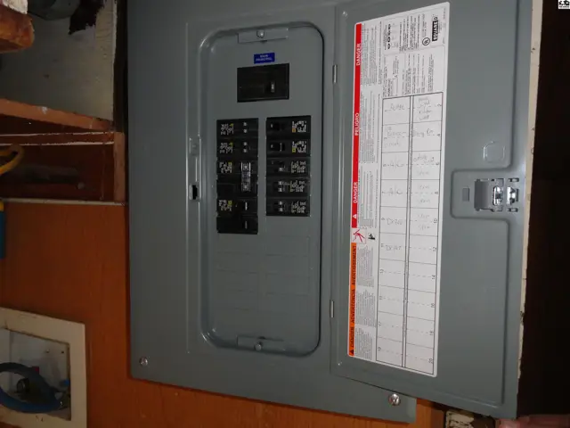Gray electrical panel with breakers and a labeled cover, mounted on an orange wall.
