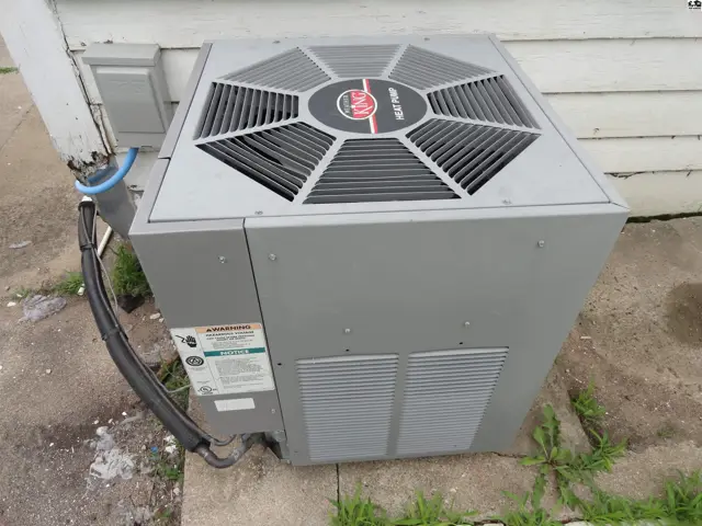 Gray outdoor heat pump unit with a black hose and electrical conduit attached.