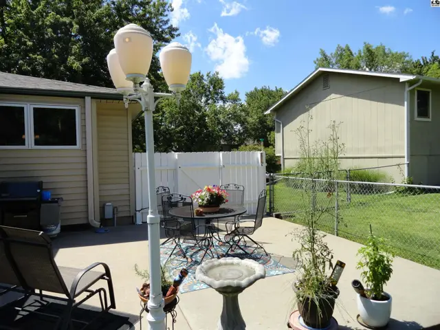 Concrete patio with outdoor dining set, grill, and landscaping under a sunny sky.