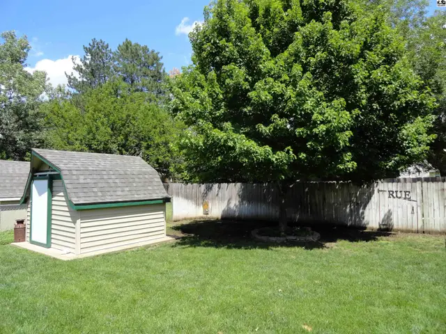 A tan shed with a green trim and shingled roof sits on a green lawn next to a wooden fence.