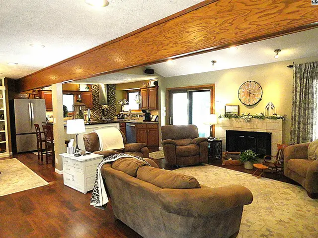 Spacious living room with plush seating, stone fireplace, and exposed wood beam ceiling, opening to a modern kitchen.