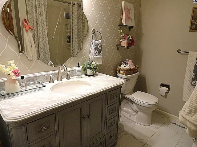 Elegant bathroom features a gray vanity with marble countertop, ornate mirror, and decorative wallpaper.