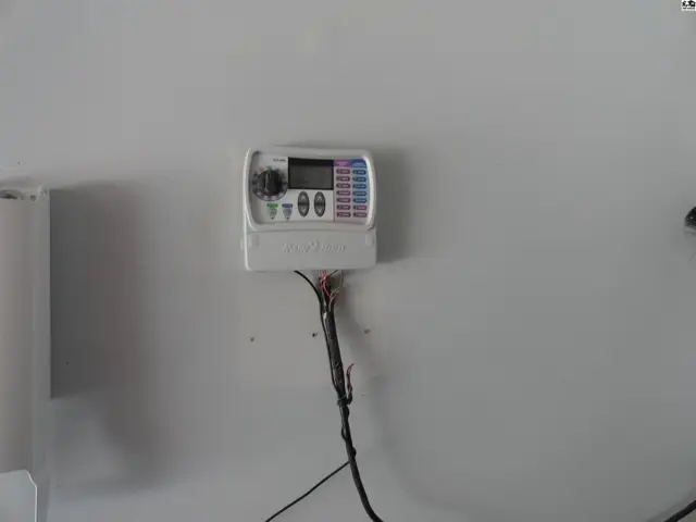 Wall-mounted irrigation controller with digital display and exposed wiring on a plain wall.