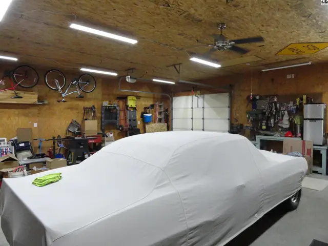 Spacious garage with wood-paneled walls, ceiling fan, and a car covered with a white tarp.