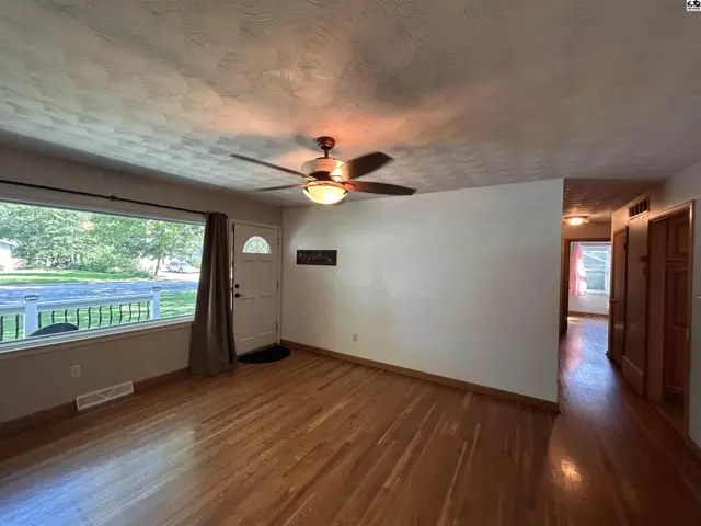 Spacious living room with hardwood floors, large window, and ceiling fan, opening to a hallway.