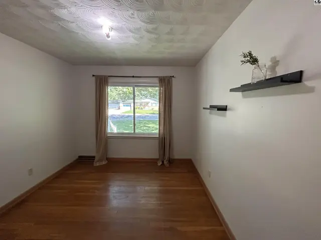 Empty room with hardwood floors, white walls, and a large window with beige curtains.