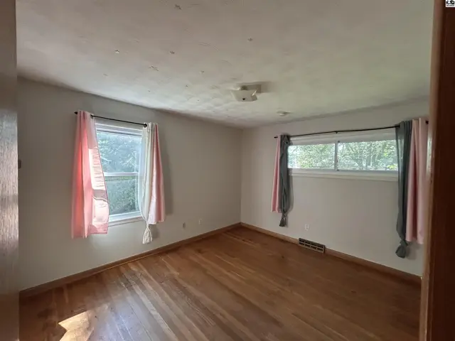 Bright bedroom with hardwood floors, two windows with pink and gray curtains, and a ceiling light fixture.