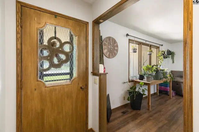 Warm wood door with decorative glass panel opens to a bright entryway with plants and wood flooring.