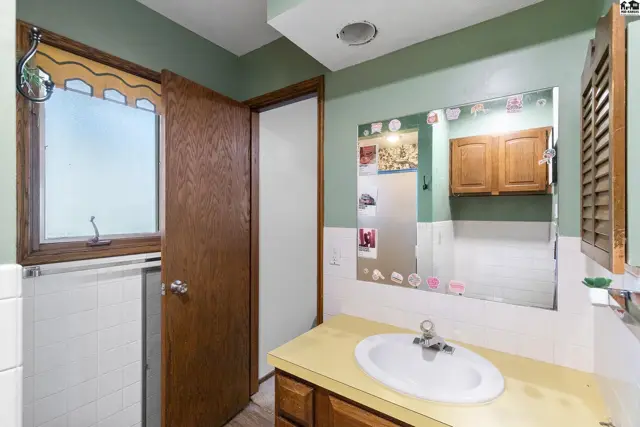 Bathroom with a light green vanity, white sink, and wood cabinets reflected in a large mirror.