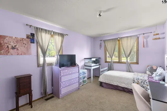 Lavender bedroom with a bed, dresser, TV, and large windows overlooking greenery.