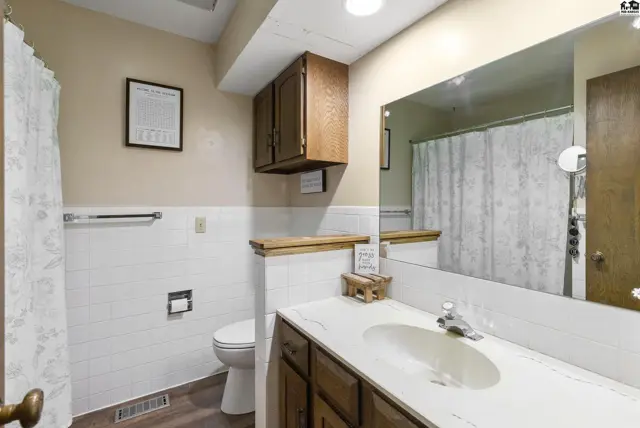 Bright bathroom with white tile, wood vanity, and a large mirror reflecting the shower curtain.