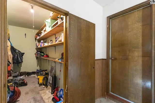 Pantry with wood shelving and storage, adjacent to a wood-paneled hallway with a closed door.