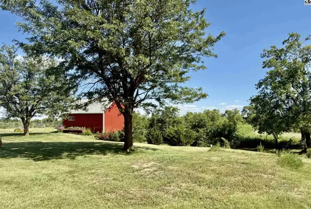 Red barn with white trim sits amidst lush green trees and rolling landscape under a clear blue sky.
