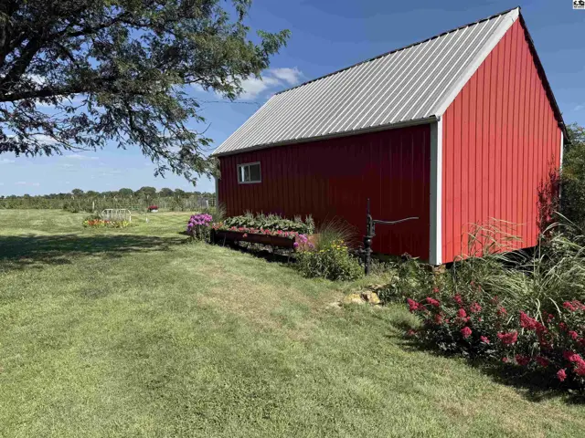 Red barn with a metal roof sits in a grassy yard with blooming flowers.