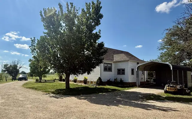 White vinyl-sided house with a carport, mature trees, and a gravel driveway under a blue sky.