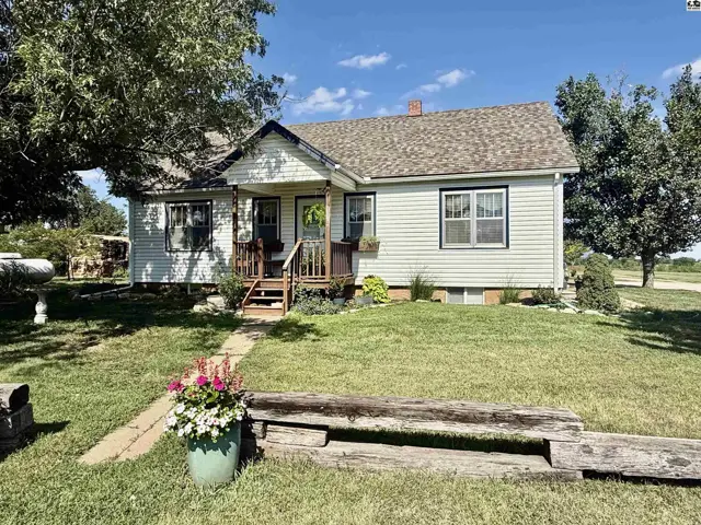 Charming white vinyl-sided farmhouse with a welcoming porch and mature trees on a sunny day.