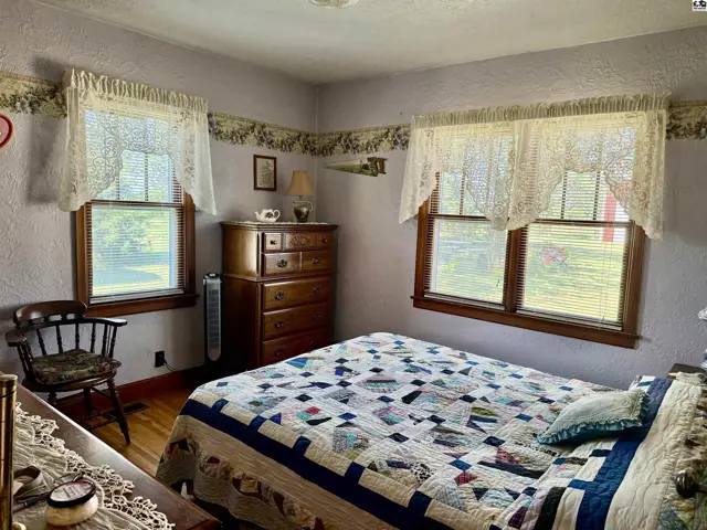 Cozy bedroom with a quilt-covered bed, wooden furniture, and lace-curtained windows.