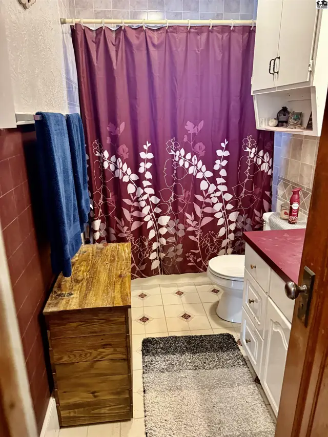 Compact bathroom with a floral shower curtain, white vanity, and wooden storage chest.