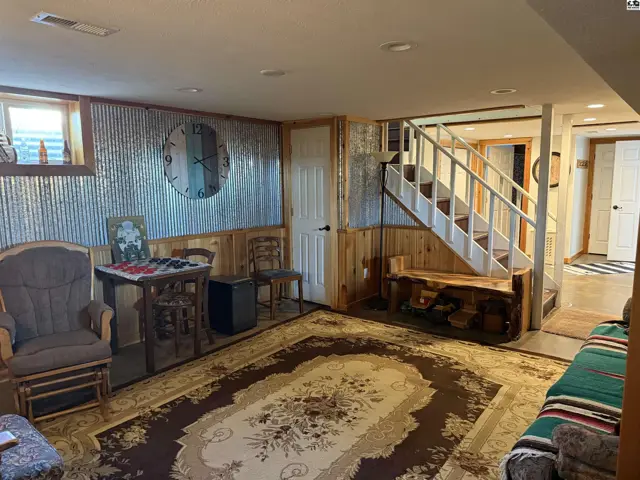 Rustic basement living area with corrugated metal walls, wood paneling, a large rug, and a staircase.