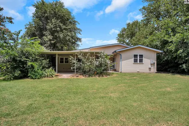 Exterior view of a beige house with a covered patio and mature trees in a grassy yard.