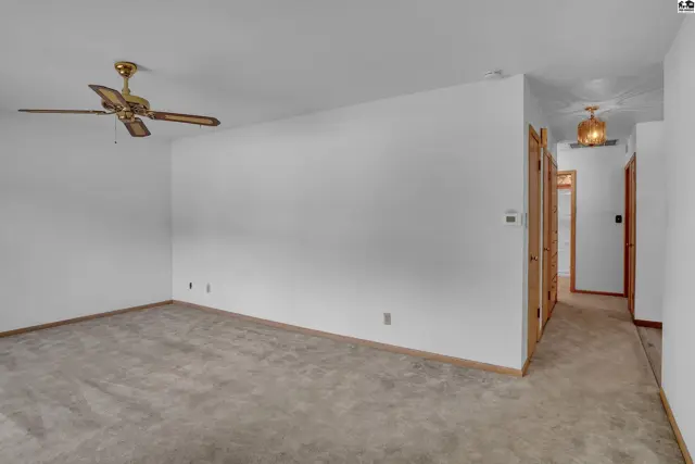 Spacious living area with neutral carpet, white walls, and a brass ceiling fan, leading to a hallway.