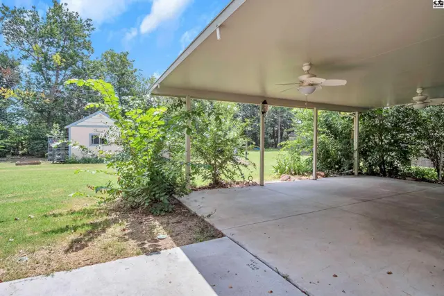 Carport with concrete floor, ceiling fans, and surrounding green lawn and trees.