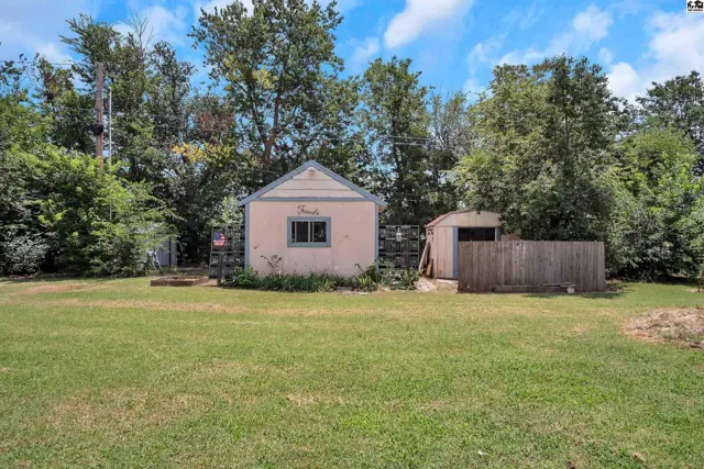 A small, light-pink cottage with a peaked roof sits in a grassy yard with mature trees.