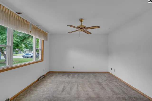 Spacious living room with large window, neutral carpet, and brass ceiling fan.