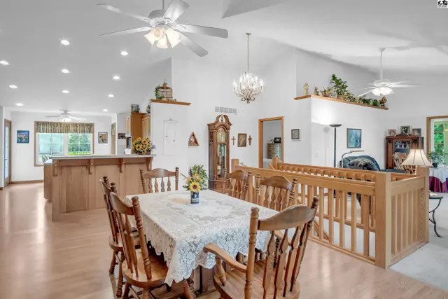 Spacious dining area with a lace-covered table, wooden chairs, and a grandfather clock, opening to a bright, open-concept living space.