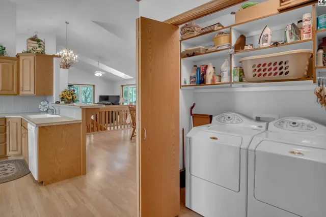 Laundry room with white washer and dryer next to a kitchen with light wood cabinets and a sink.