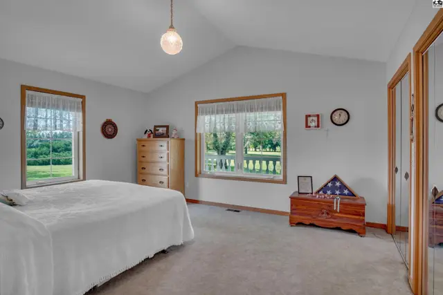 Spacious bedroom with vaulted ceiling, large windows, carpet, and wooden furniture.
