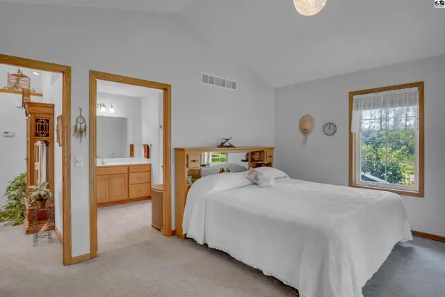 Spacious bedroom with a white bed, mirrored headboard, and a view of greenery through the window.