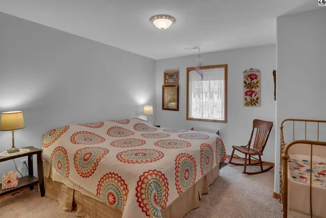 Cozy bedroom with a large bed, patterned quilt, rocking chair, and neutral carpet.