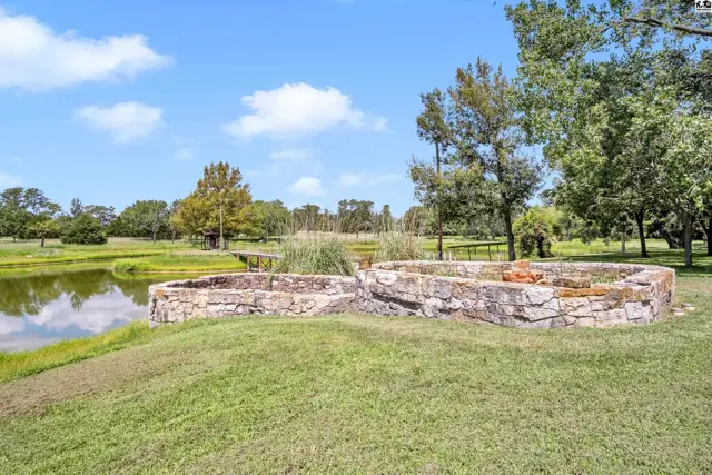 Stone fire pit and pond on a grassy landscape with mature trees under a blue sky.