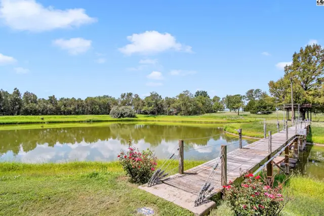 Wooden dock with railing leads to a serene pond surrounded by lush green trees and vibrant red flowers.