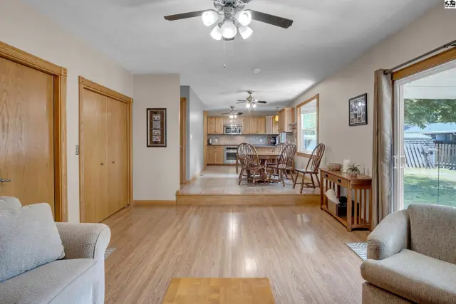 Open living area with light wood floors, adjacent kitchen, and large windows overlooking the yard.