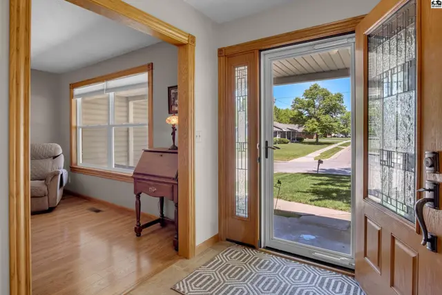 Warm wood door with decorative glass opens to a bright entryway with a view of a tree-lined street.