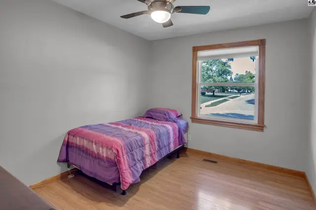 Bedroom with light wood floors, gray walls, a ceiling fan, and a large window overlooking a street.