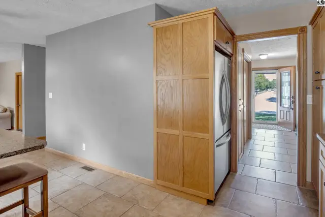 Kitchen with light wood cabinetry, stainless steel refrigerator, and tiled flooring leading to a hallway.
