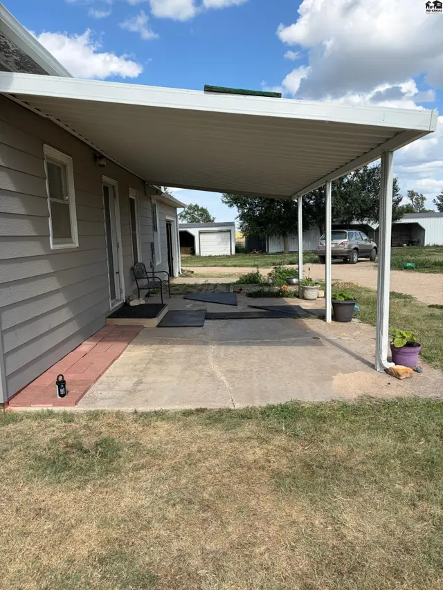 Covered concrete patio with gray siding and white trim, featuring outdoor seating and potted plants.