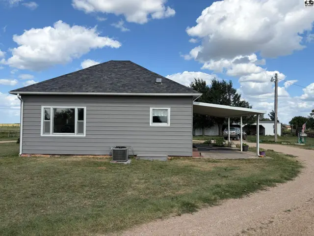 Gray siding house with a white carport and large window under a cloudy blue sky.