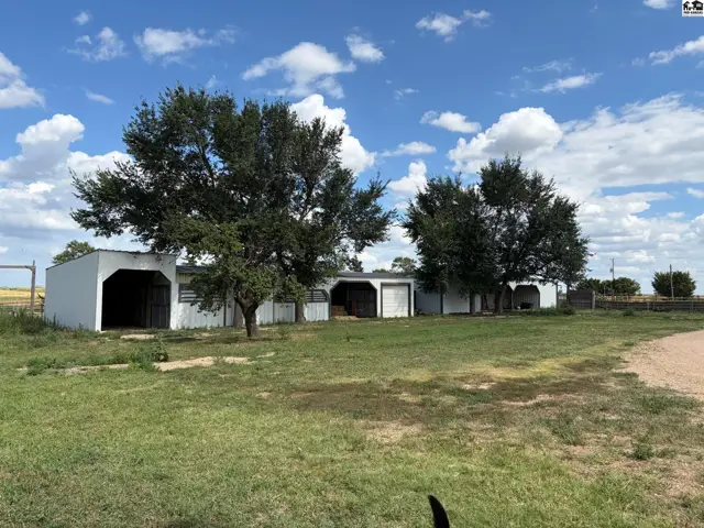 White stables with open stalls and mature trees under a cloudy blue sky.