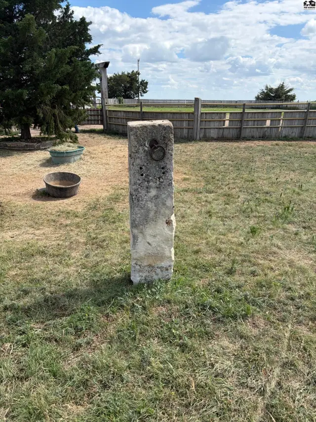 Stone hitching post with metal ring stands in a grassy yard with a wooden fence.